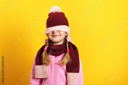 Cheerful little girl in a scarf and a hat on a yellow background. The child hides his eyes under a hat