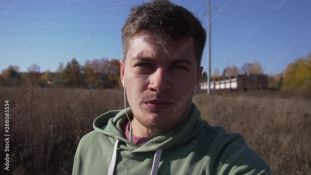 Cute caucasian young male, wearing light-green hoody is slowly walking alone in country side on narrow road covered with green high grass along empty wheat fields on clear day with blue sky. Cute caucasian young male, wearing light-green hoody is slowly walking alone in country side on narrow road covered with green high grass along empty wheat fields on clear day with blue sky.