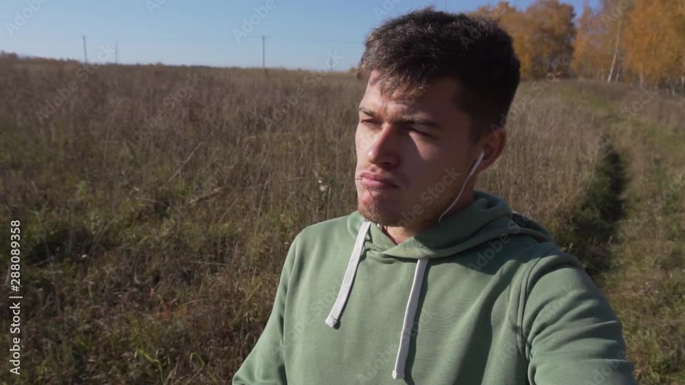 Attractive caucasian young man with thin stalk in mouth, wearing light-green hoody, is slowly walking alone in countryside along empty field on clear autumn day with blue sky. Attractive caucasian young man with thin stalk in mouth, wearing light-green hoody, is slowly walking alone in countryside along empty field on clear autumn day with blue sky.