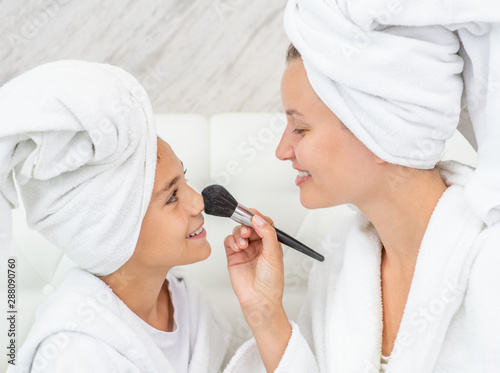 Happy family at home. Smiling mom applying powder on nose her little daughter using a brush. Woman and girl are in bathrobes and with towels on their heads