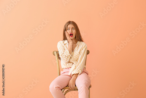 Shocked young woman sitting on chair against color background