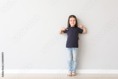 Fototapeta Naklejka Na Ścianę i Meble -  Cute little girl pointing at her t-shirt near light wall