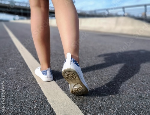 In the frame, the legs of a young girl in white sports shoes, which is walking along a road marking on an asphalt path.