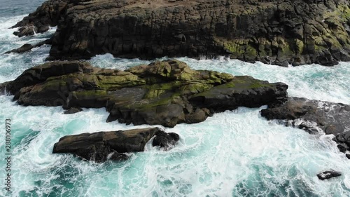 Drone looks down over sea rocks as waves crash into it.
