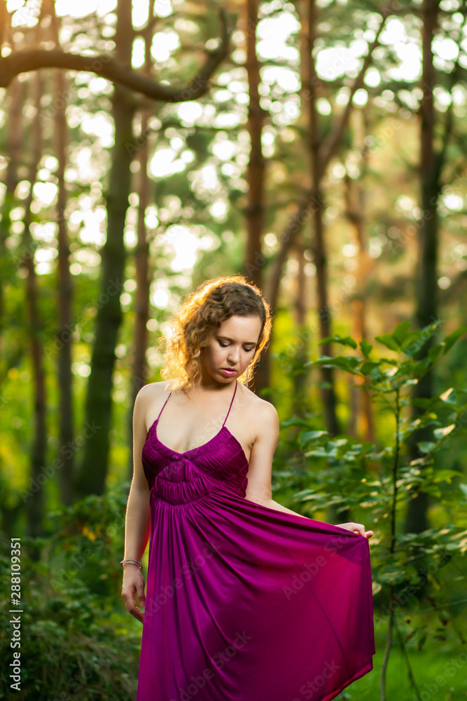 Beautiful curly hair young woman wearing elegant purple dress standing in the middle of forest with rays of sunlight during sunset Beautiful curly hair young woman wearing elegant purple dress standing in the middle of forest with rays of sunlight during sunset