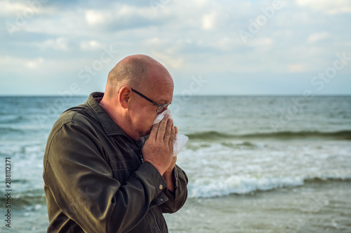 A man stands by the sea and uses a handkerchief, he has an allergy or severe flu. Concept: Runny nose or protection against infections © andre