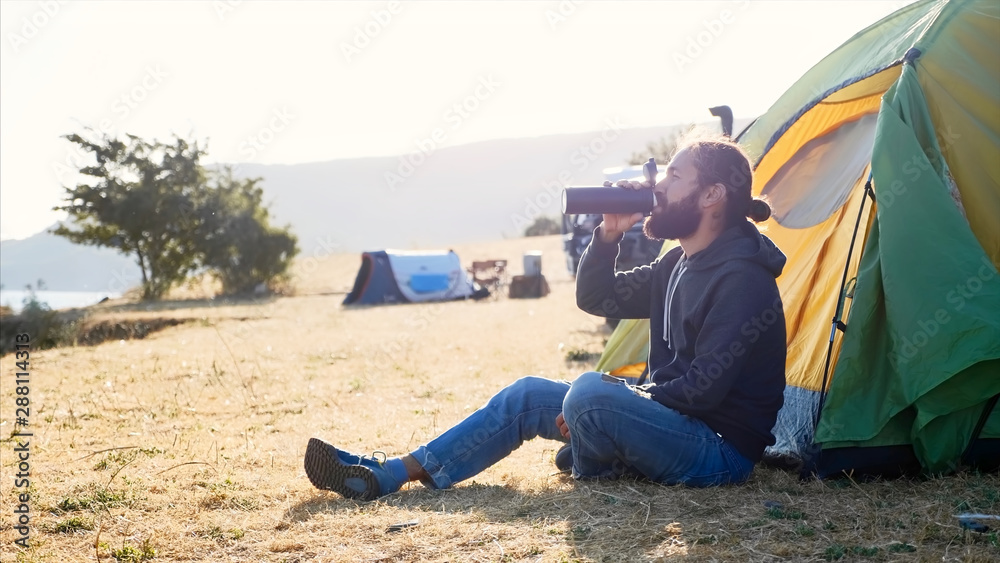 Young bearded hipster man sits on the ground next to touristic tent at expedition campsite on lake shore and drinking tea or coffee from thermos bottle at morning. Side view with backlit