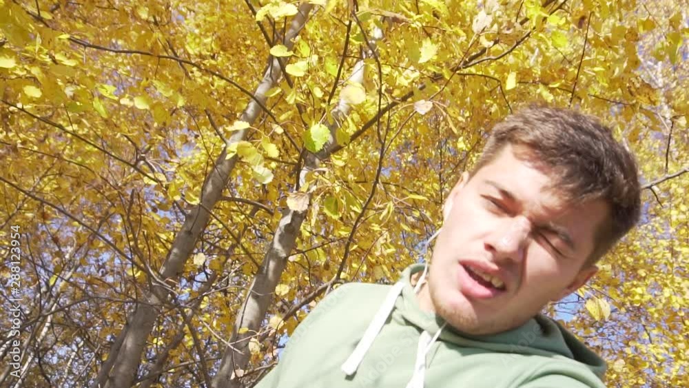 View of caucasian young nice-looking guy, wearing light-green hoody is standing under thin trees with yellow leaves somewhere in countryside on sunny Autumn day with blue sky. View of caucasian young nice-looking guy, wearing light-green hoody is standing under thin trees with yellow leaves somewhere in countryside on sunny Autumn day with blue sky.