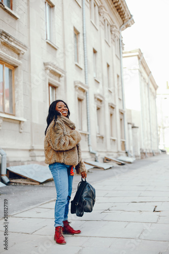 A beautiful and stylish black girl with long dark hair dressed in a brown coat and shirt wallking in a summer city © hetmanstock2