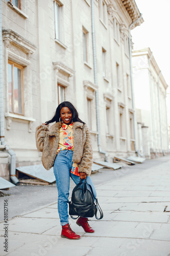 A beautiful and stylish black girl with long dark hair dressed in a brown coat and shirt wallking in a summer city