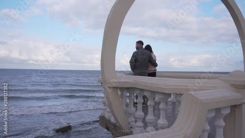 Man and woman are standing on white, empty viewing resort platform and looking at beautiful coastline with big waves slowly falling on shores with stones and rocks on windy summer day. photo