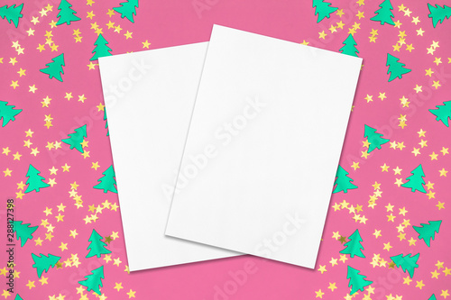Two empty white rectangle poster mockups lying diagonally on top of each other with soft shadows on pink background with metallic foil christmas trees and gold stars confetti. Flat lay, top view