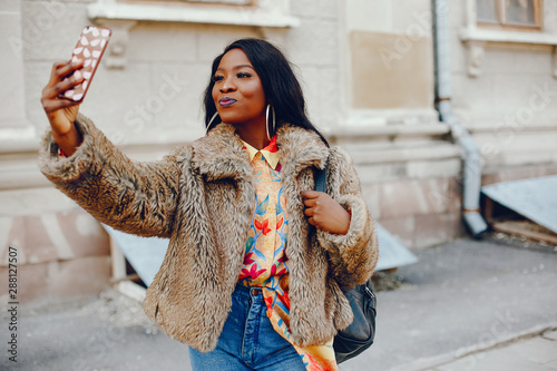 A beautiful and stylish black girl with long dark hair dressed in a brown coat and shirt wallking in a summer city with phone in case