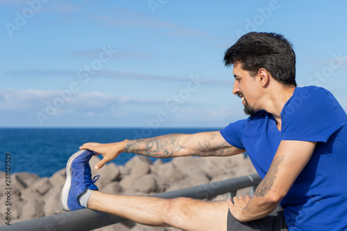 Fitness Runner smiling while stretching his leg in a fence in front of the sea. Athletic boy balance after training in the seaside