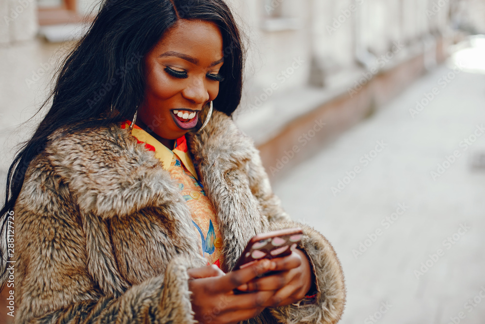 A beautiful and stylish black girl with long dark hair dressed in a brown coat and shirt wallking in a summer city with phone in case A beautiful and stylish black girl with long dark hair dressed in a brown coat and shirt wallking in a summer city with phone in case