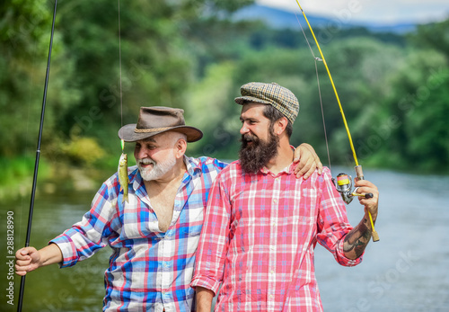 Hugs Are Fully Returnable. summer weekend. mature men fisher. male friendship. family bonding. two happy fisherman with fishing rods. hobby and sport activity. Trout bait. father and son fishing