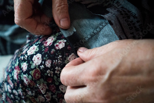 Macro elderly woman hands sewing on fabric, needle. Sew pants by old woman hand, seamstress at work with cloth fabric. Grandmother sews. Close up shot. Selective focus