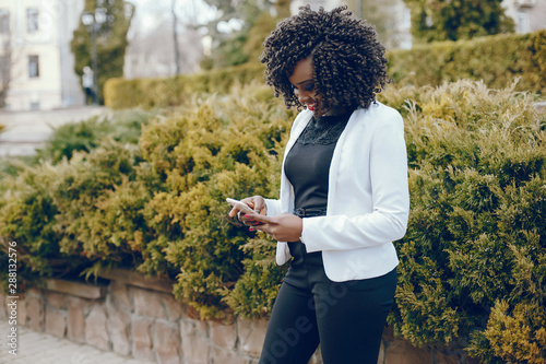 elegant and stylish dark-skinned girl with curly hair and in a white jacket walking around the summer city and using the phone
