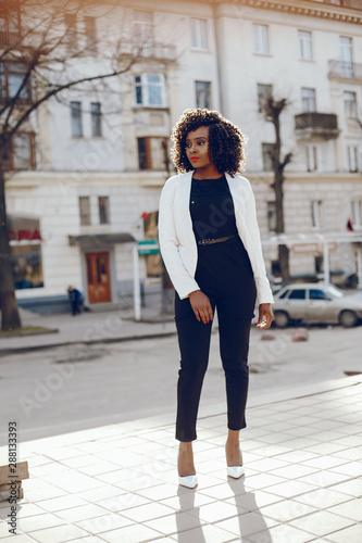 elegant and stylish dark-skinned girl with curly hair in a white jacket walking around the summer city