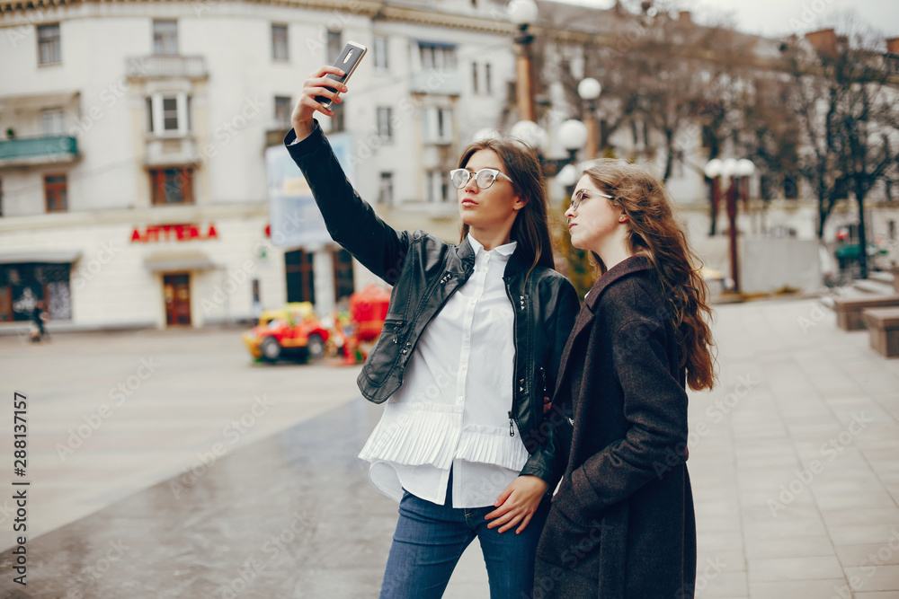 a beautiful stylish young girl with long curly hair and a long coat walking in the autumn city with her girlfriend in a black leather jacket and glasses and they uses the phone