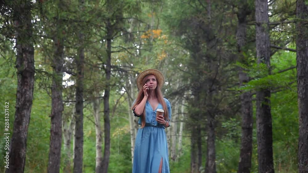Young beautiful woman with long hair in straw hat in blue dress walking in a forest, talking by mobile phone, holding a cup of coffee Young beautiful woman with long hair in straw hat in blue dress walking in a forest, talking by mobile phone, holding a cup of coffee