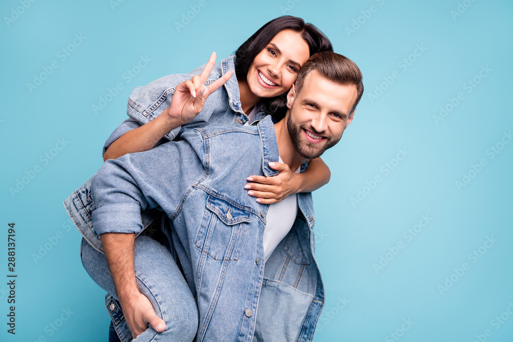 Portrait of cute man carrying his sweetheart making v-signs wearing denim jeans jacket isolated over blue background Portrait of cute man carrying his sweetheart making v-signs wearing denim jeans jacket isolated over blue background