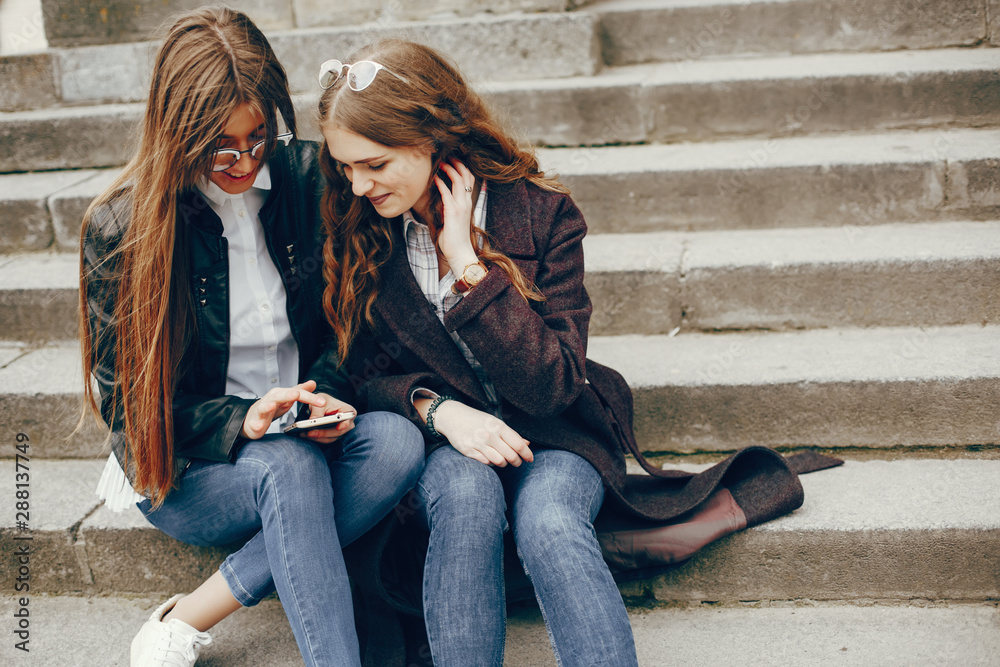 a beautiful stylish young girl with long curly hair and a long coat sitting in the autumn city with her girlfriend in a black leather jacket and glasses and they uses the phone a beautiful stylish young girl with long curly hair and a long coat sitting in the autumn city with her girlfriend in a black leather jacket and glasses and they uses the phone
