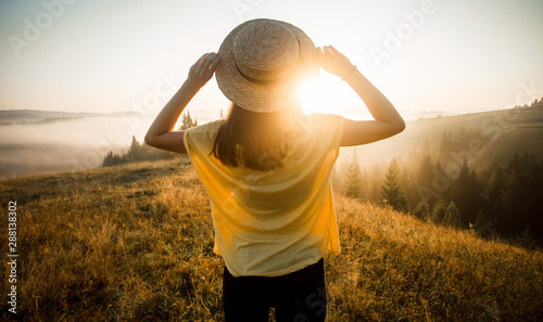Back view of woman in straw hat stay outdoor under sunlight of sunrise with beautiful view of forest mountains