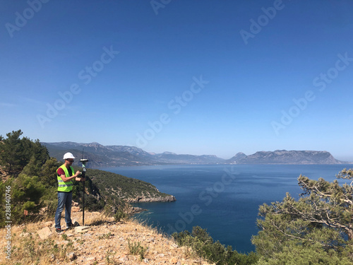 Fototapeta Naklejka Na Ścianę i Meble -  Geodetic engineer surveyor in white hard hat doing measurements with GNSS satellite receiver at the hill top