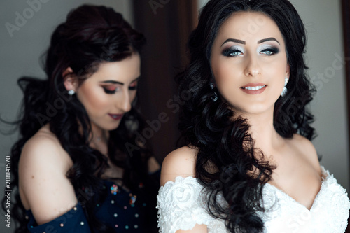  Photo of a friend of honor helping the bride with her dress.  Bridesmaid help bride at her home. Morning of beaufiful bride. Girl's hepl bride look wonderful. Makeup and hairstyle. © Tetiana Moish