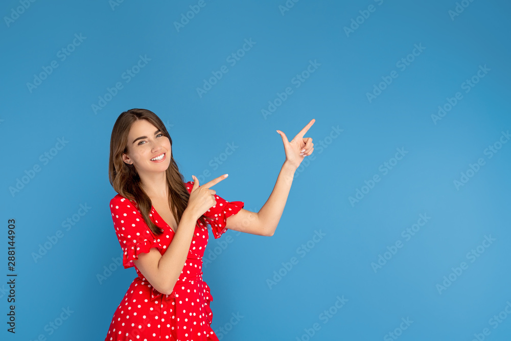 Beautiful young woman with bright smile in red dress pointing fingers up over blue background. Beautiful young woman with bright smile in red dress pointing fingers up over blue background.