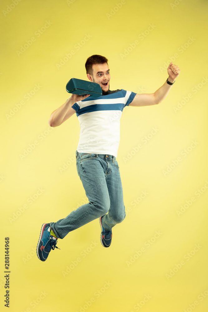 Sky sound. Full length portrait of happy jumping man with gadgets on yellow background. Modern tech, freedom of choices concept, emotions concept. Using portable speaker like superhero in flight. Sky sound. Full length portrait of happy jumping man with gadgets on yellow background. Modern tech, freedom of choices concept, emotions concept. Using portable speaker like superhero in flight.