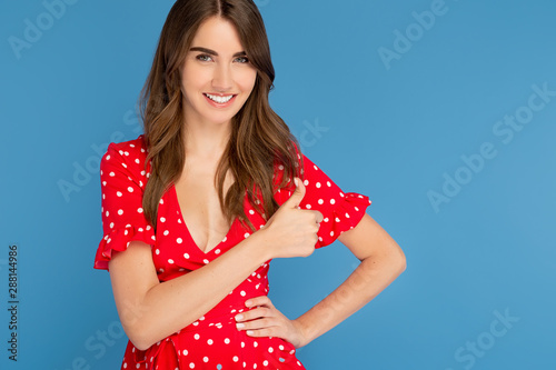 Cheerful young woman with bright smile in red dress showing OK gesture over blue background. © Screaghin