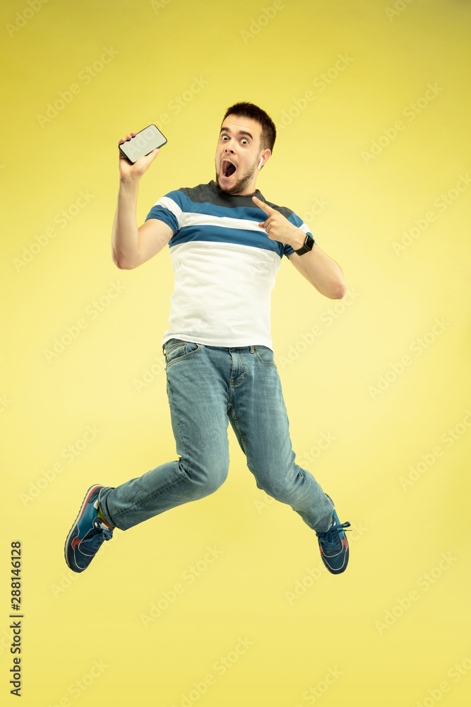 Communication. Full length portrait of happy jumping man with gadgets on yellow background. Modern tech, freedom of choices concept, emotions concept. Using smartphone for selfie or videocall in