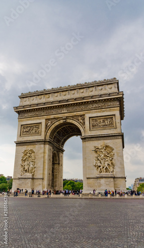 Nice portrait view of the famous monument Arc de Triomphe in Paris, seen from the east with the two sculptures Le Départ and Le Triomphe on a cloudy, rainy day in front of the cobblestone road. © H-AB Photography
