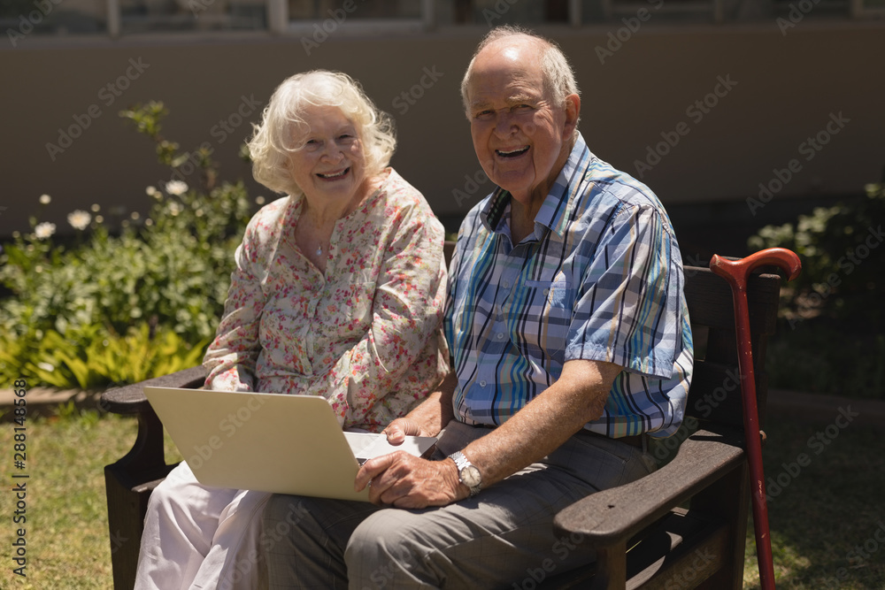 Front view of senior couple with laptop looking at camera in garden Front view of senior couple with laptop looking at camera in garden