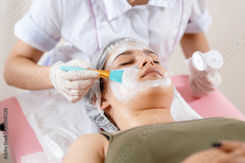 Cosmetology specialist applying facial mask using brush  making skin hydrated and healthy. Attractive woman relaxing. Beautician at work. Hands of cosmetology