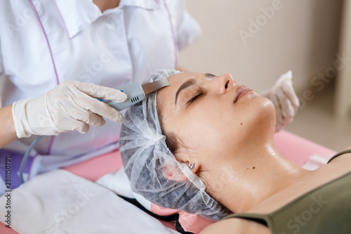 Beauty Specialist Makes Ultrasonic Peeling For Female Client's Face. Cosmetologist doing procedure of cleaning face with ultrasonic scrubber. In beauty salon