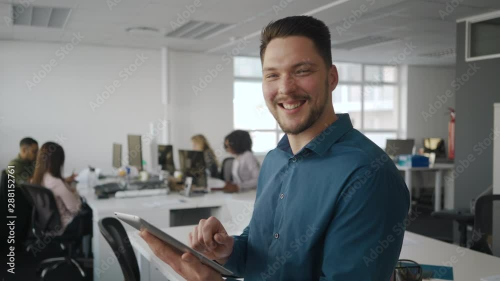 Young smiling businessman using tablet computer looking and smiling at camera and his creative team working at background Young smiling businessman using tablet computer looking and smiling at camera and his creative team working at background