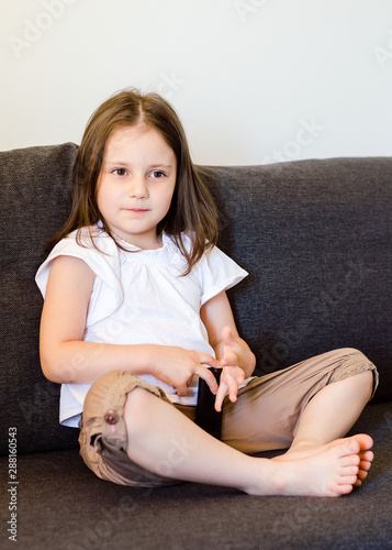 A 4-year-old girl sits on a gray sofa with a TV remote control and watches TV