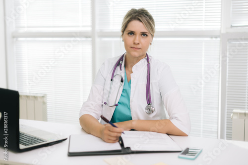 Portrait of young woman doctor in hospital office with laptop taking patient. Female therapist sits at the table in front of the camera and looking at camera