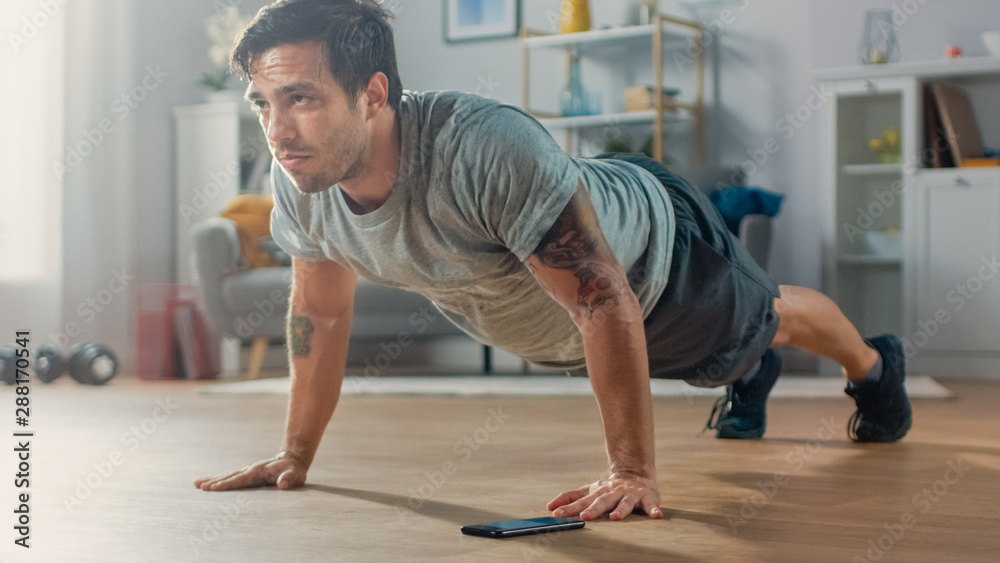 Athletic Fit Man in T-shirt and Shorts is Doing Push Up Exercises While Using a Stopwatch on His Phone. He is Training at Home in His Living Room with Minimalistic Interior.