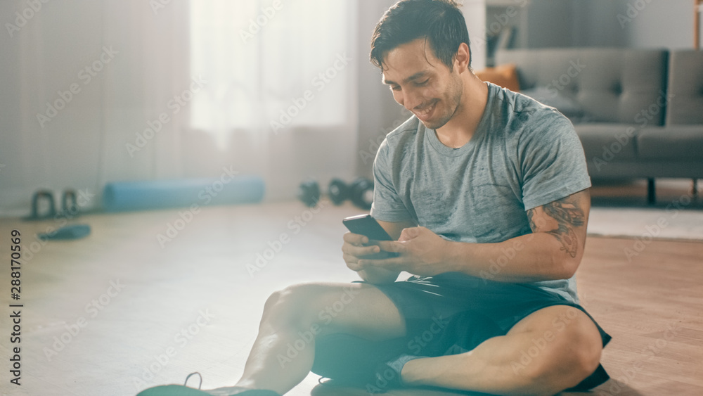 Obraz premium Happy Strong Athletic Fit Man in T-shirt and Shorts is Using a Mobile Phone After Morning Exercises at Home in His Spacious and Bright Living Room with Minimalistic Interior.