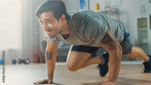 Muscular Athletic Fit Man in T-shirt and Shorts is Doing Mountain Climber Exercises While Using a Stopwatch on His Phone. He is Training at Home in His Bright Living Room with Minimalistic Interior.