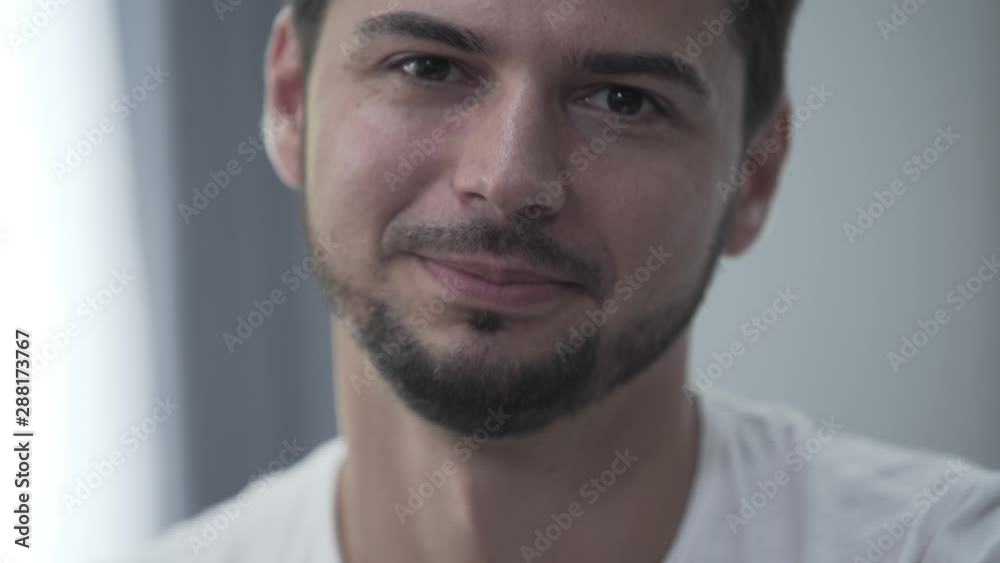 Portrait of handsome smiling man in white t-shirt sitting at home smiling while looking at the camera. Relaxing indoors. Confident bearded guy in the light room Portrait of handsome smiling man in white t-shirt sitting at home smiling while looking at the camera. Relaxing indoors. Confident bearded guy in the light room