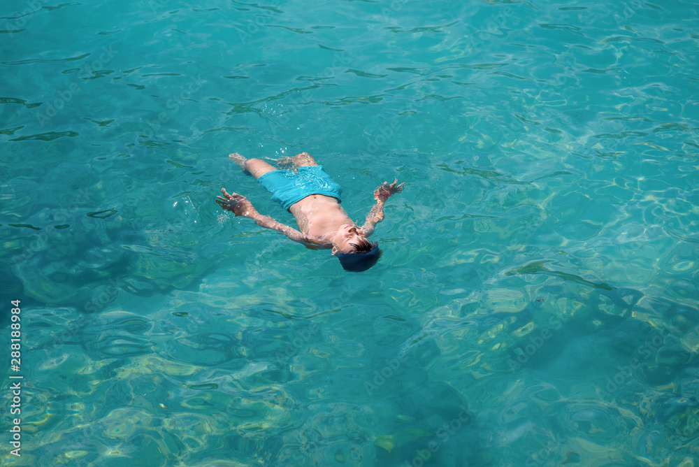 Cute European boy in blue swim pants floating in the transparent blue sea water. He is enjoying his summer holidays in Spain.
