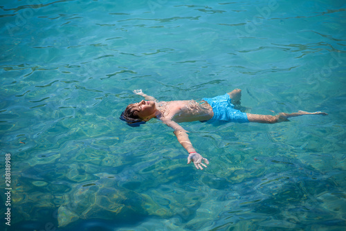 Cute European boy in blue swim pants floating in the transparent blue sea water. He is enjoying his summer holidays in Spain.