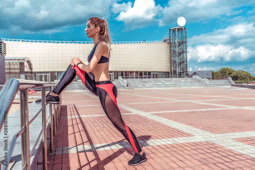 Athletic girl in summer city, does a warm-up of legs and knee, quadriceps, before fitness training run and morning. Sportswear. Leggings. Top. Sneakers. Clouds tile background, free space for text.