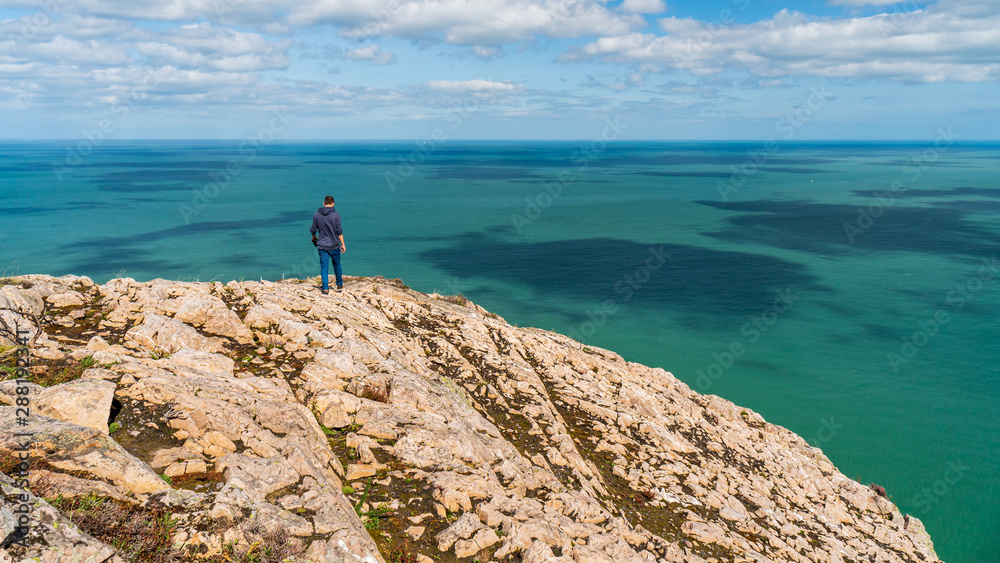 Male hiker walking towards the sea on a rocky mountain peak to admire the view.  Rugged cliffs and turquoise sea landscape.
