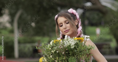 Stock footage of beautiful smiling young woman in summer dress with pink kanekalons in braids smelling wildflower bouquet in the park on summer day. photo